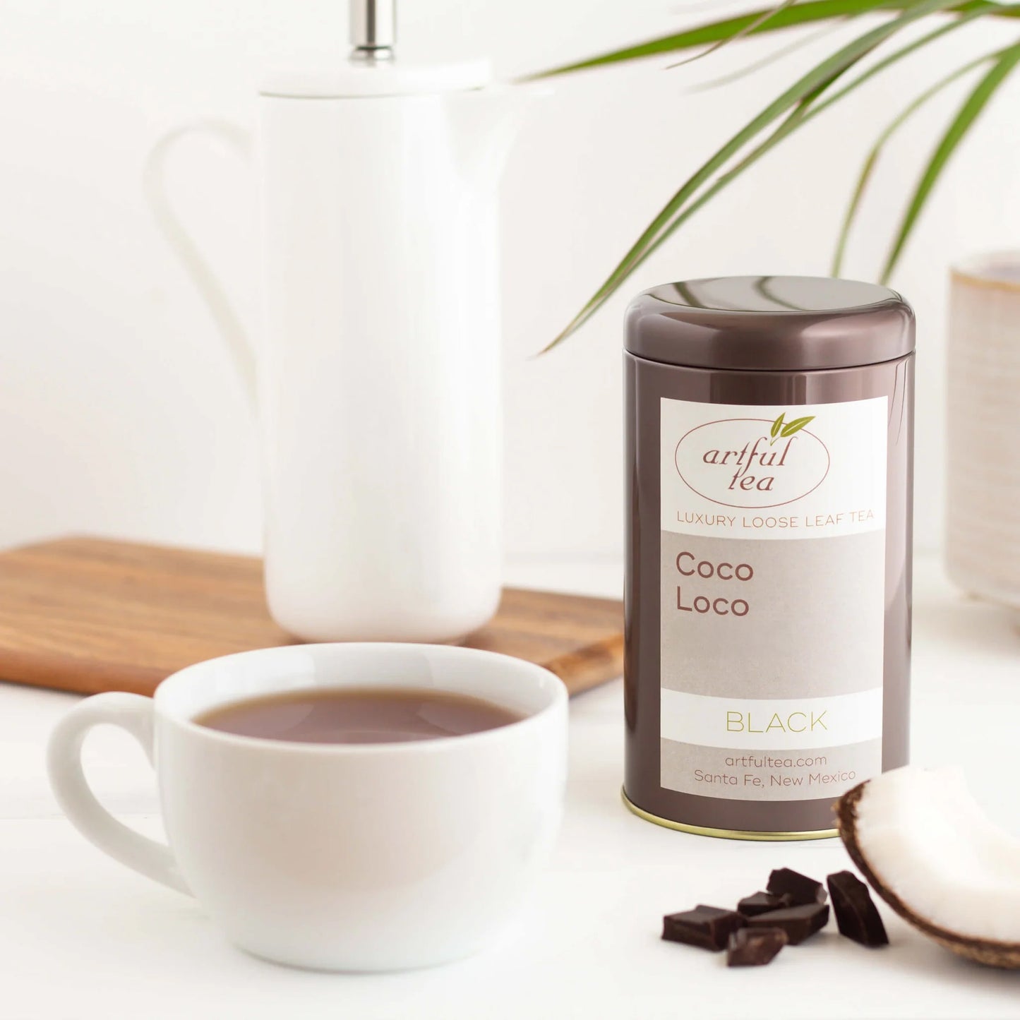 Coco Loco Black Tea shown packaged in a brown tin next to a white mug full of brewed tea with ingredients and a tall white teapot nearby.