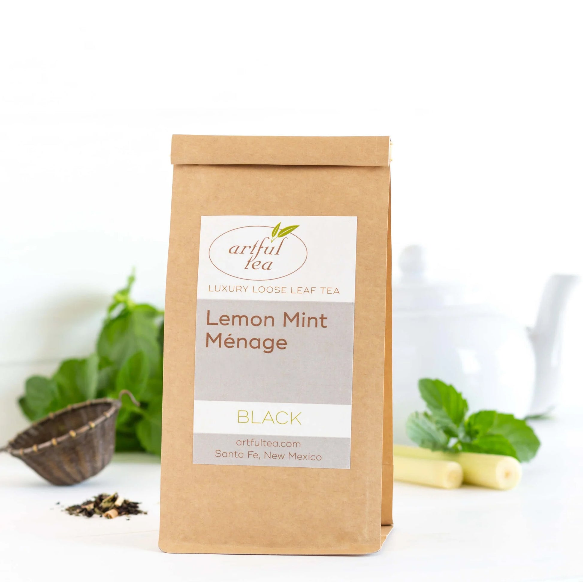Lemon Mint Ménage Black Tea shown packaged in a brown Kraft bag with ingredients nearby and a white teapot in the background.
