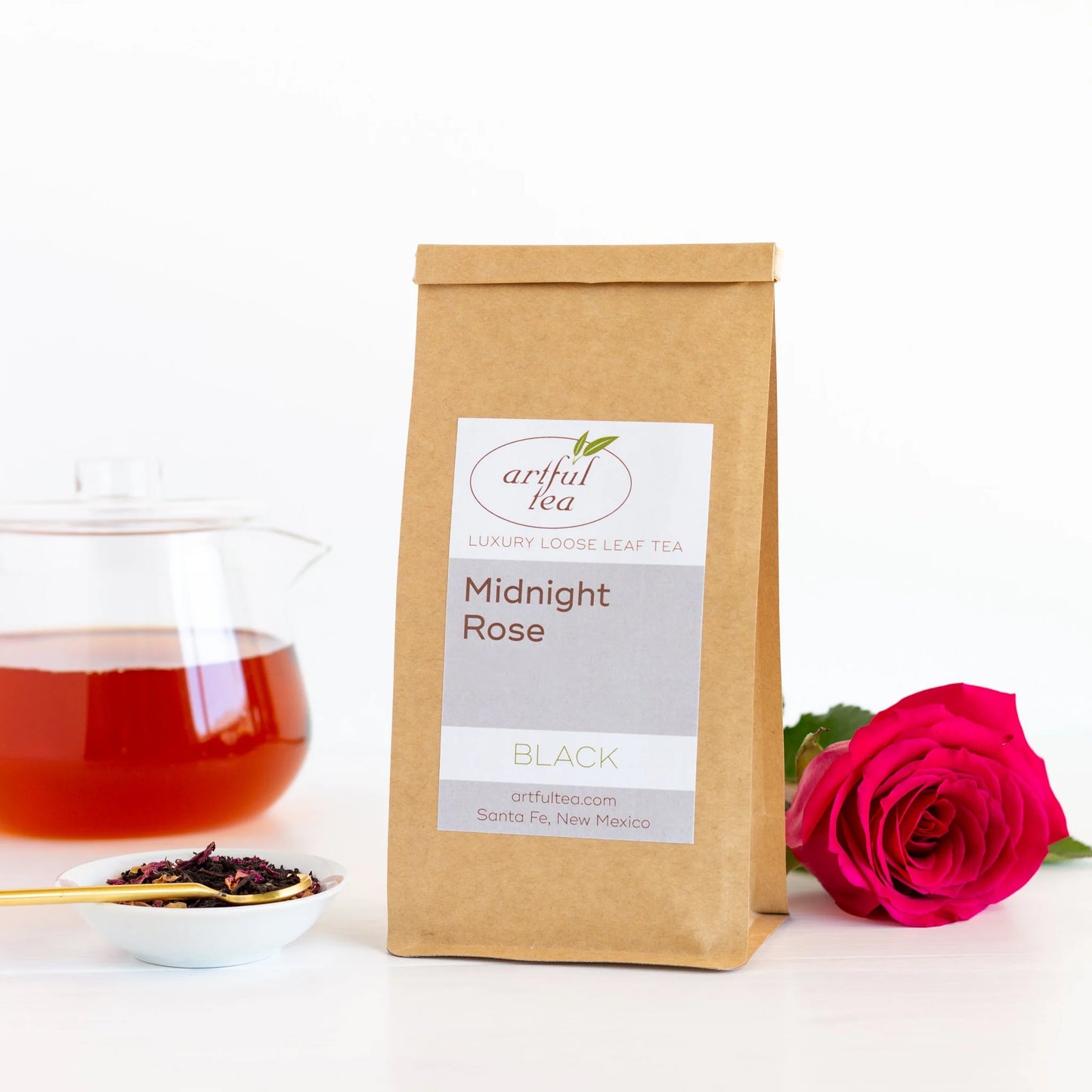 Midnight Rose Black Tea shown packaged in a brown Kraft bag next to a dark pink rose and a bowl of loose tea leaves with a gold spoon in it In the background is a clear teapot full of brewed tea.