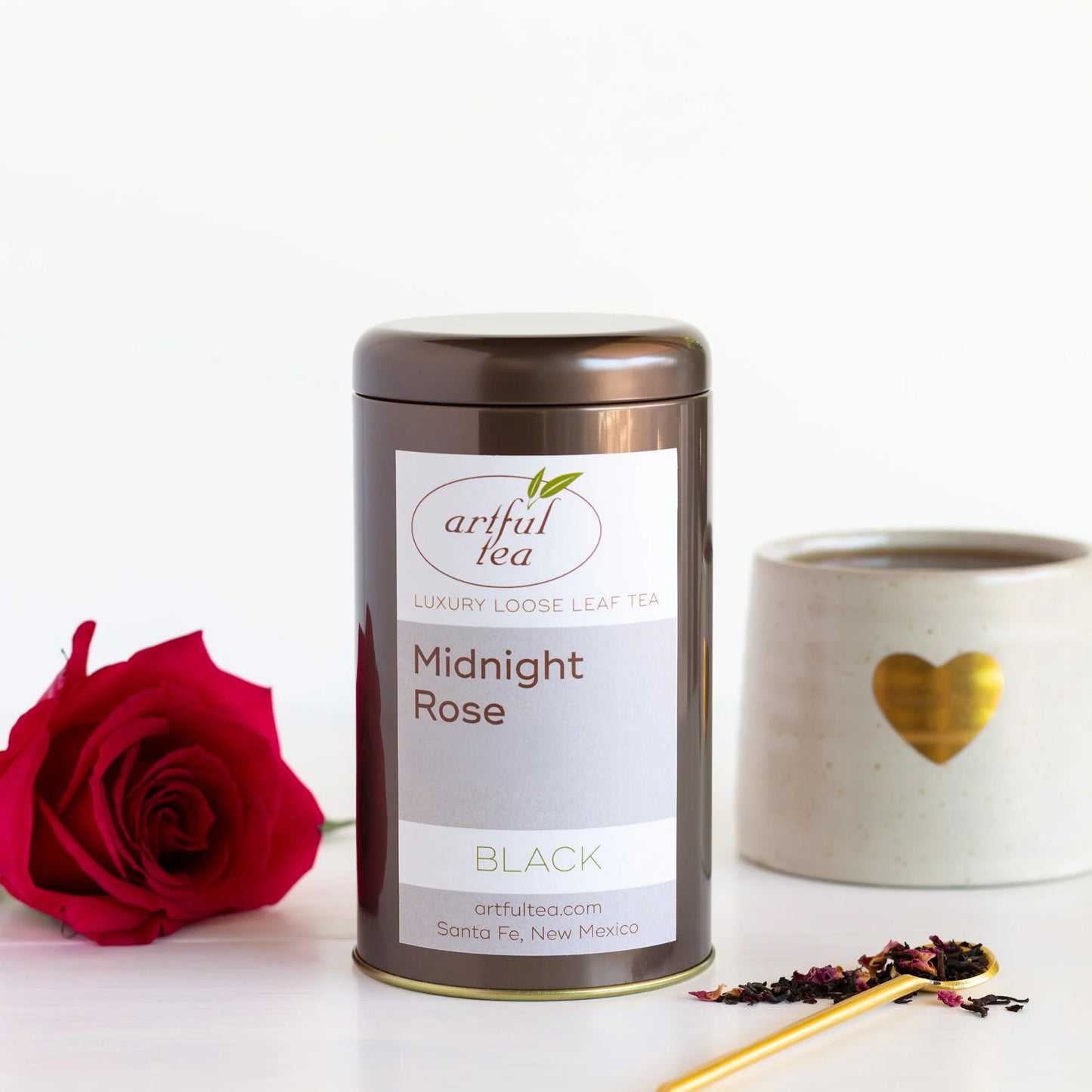 Midnight Rose Black Tea shown packaged in a brown tin between a red rose and a gold spoon full of loose tea leaves. In the background is a mug with a gold heart full of brewed tea.
