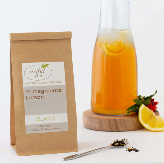 Pomegranate Lemon Black Tea shown packaged in a brown Kraft bag with a silver spoon full of loose tea leaves next to it. In the background is a large bottle of tea on a wooden coaster with ingredients nearby.