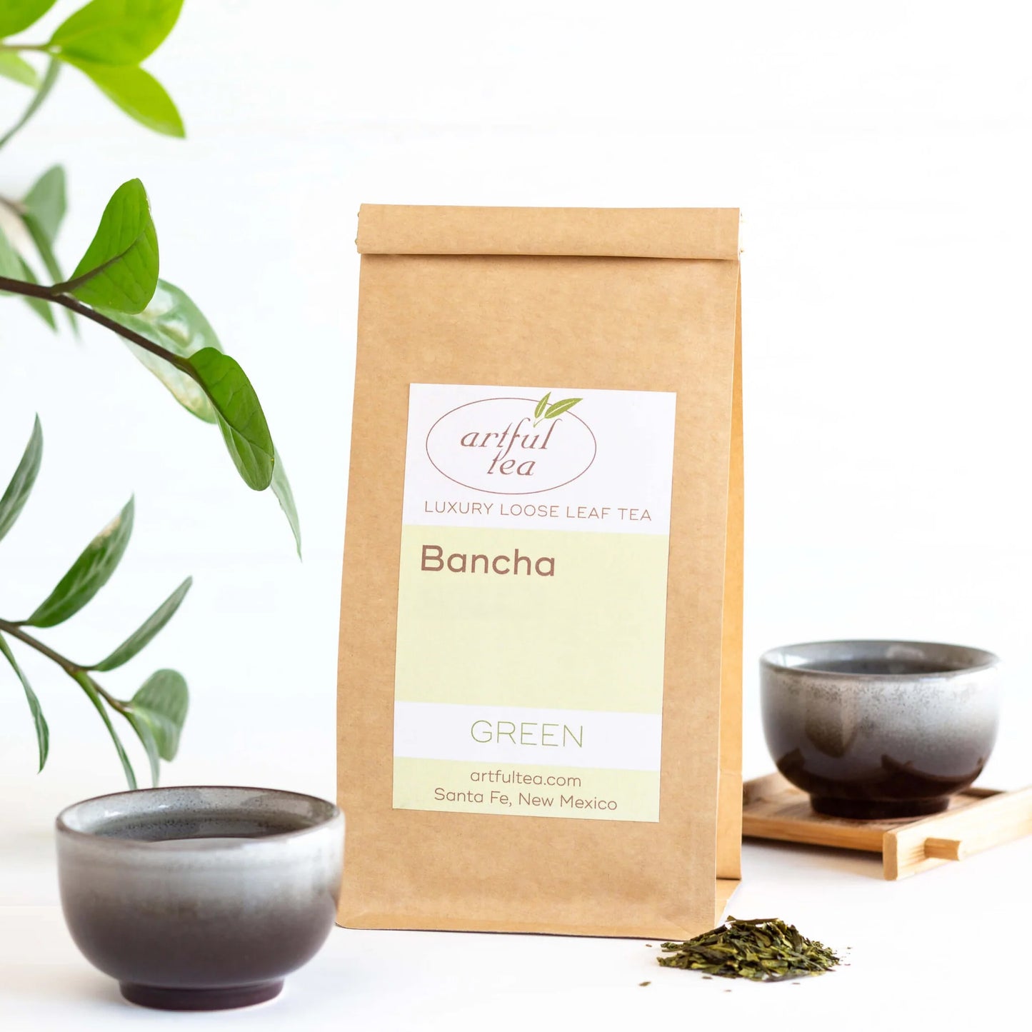 Bancha Green Tea shown packaged in a brown Kraft bag next to two teacups with loose tea leaves beside it.