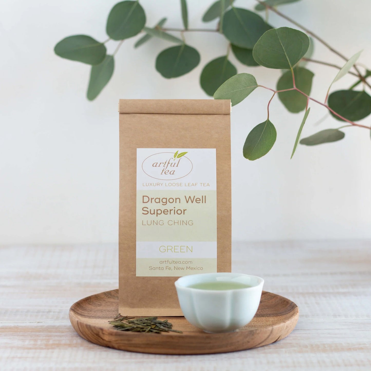 Dragon Well Superior Green Tea shown packaged in a brown Kraft bag next to a white cup of brewed tea and loose tea leaves on a circular, wooden tray.
