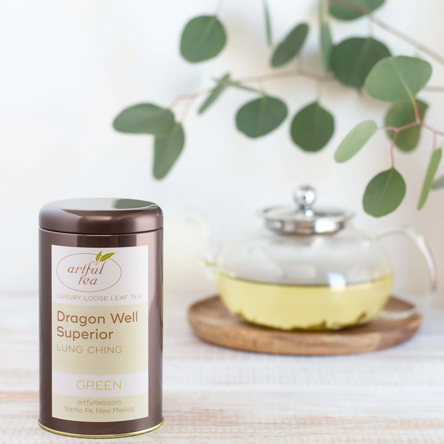 Dragon Well Superior Green Tea shown packaged in a brown tin with a clear tea pot full of brewed tea in the background.