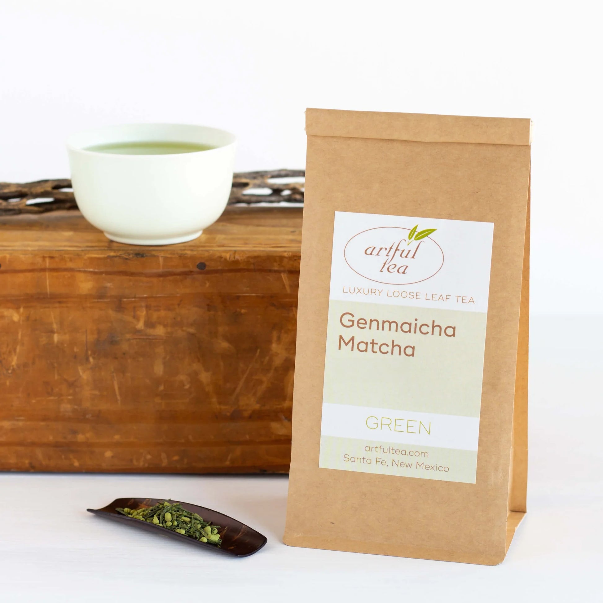 Genmaicha Matcha Green Tea shown packaged in a brown Kraft bag with a cherry wood scoop full of tea leaves beside it. In the background is a white cup of brewed tea on a wooden block.