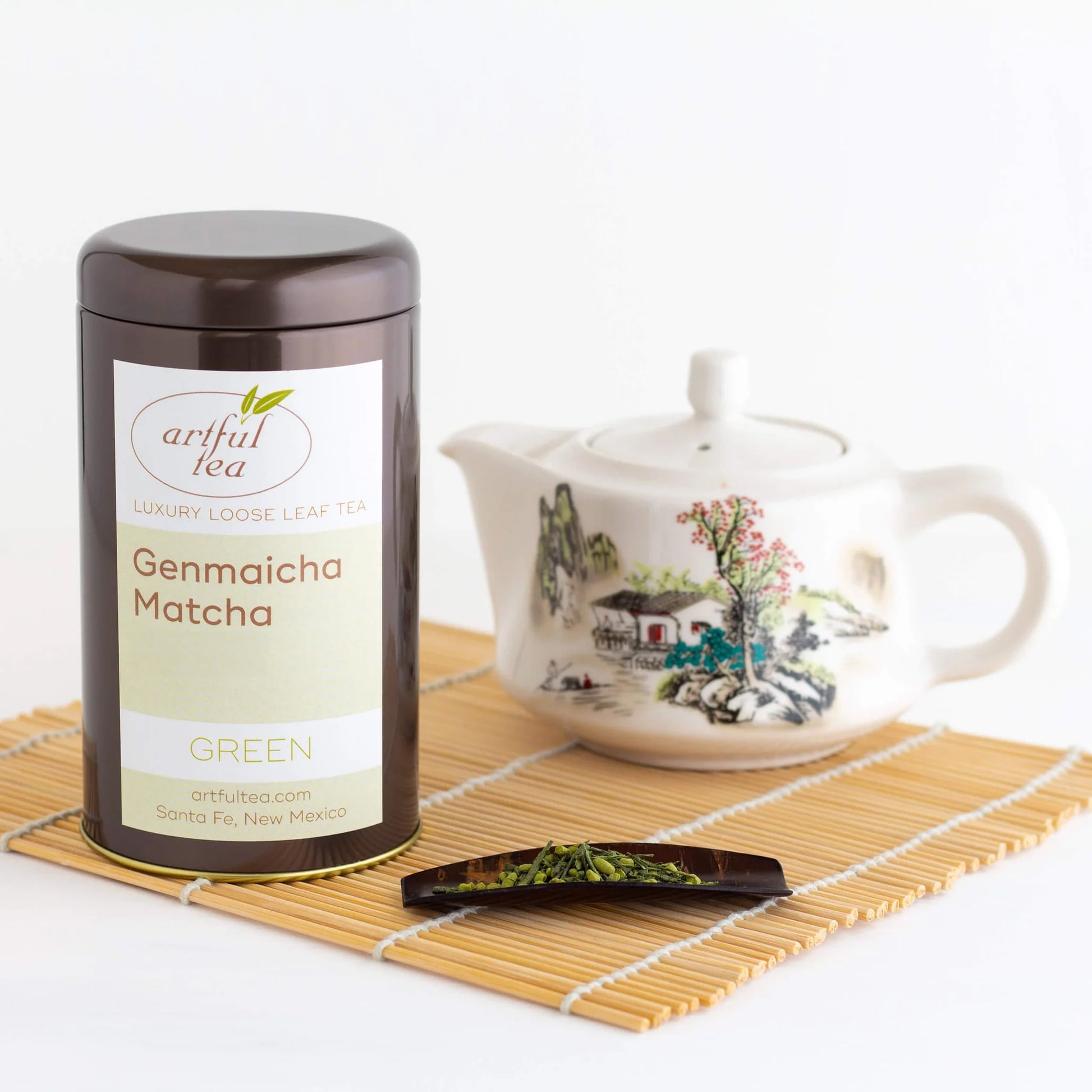 Genmaicha Matcha Green Tea shown packaged in a brown tin beside a white decorated teapot and a cherry wood scoop with tea leaves on a bamboo coaster.