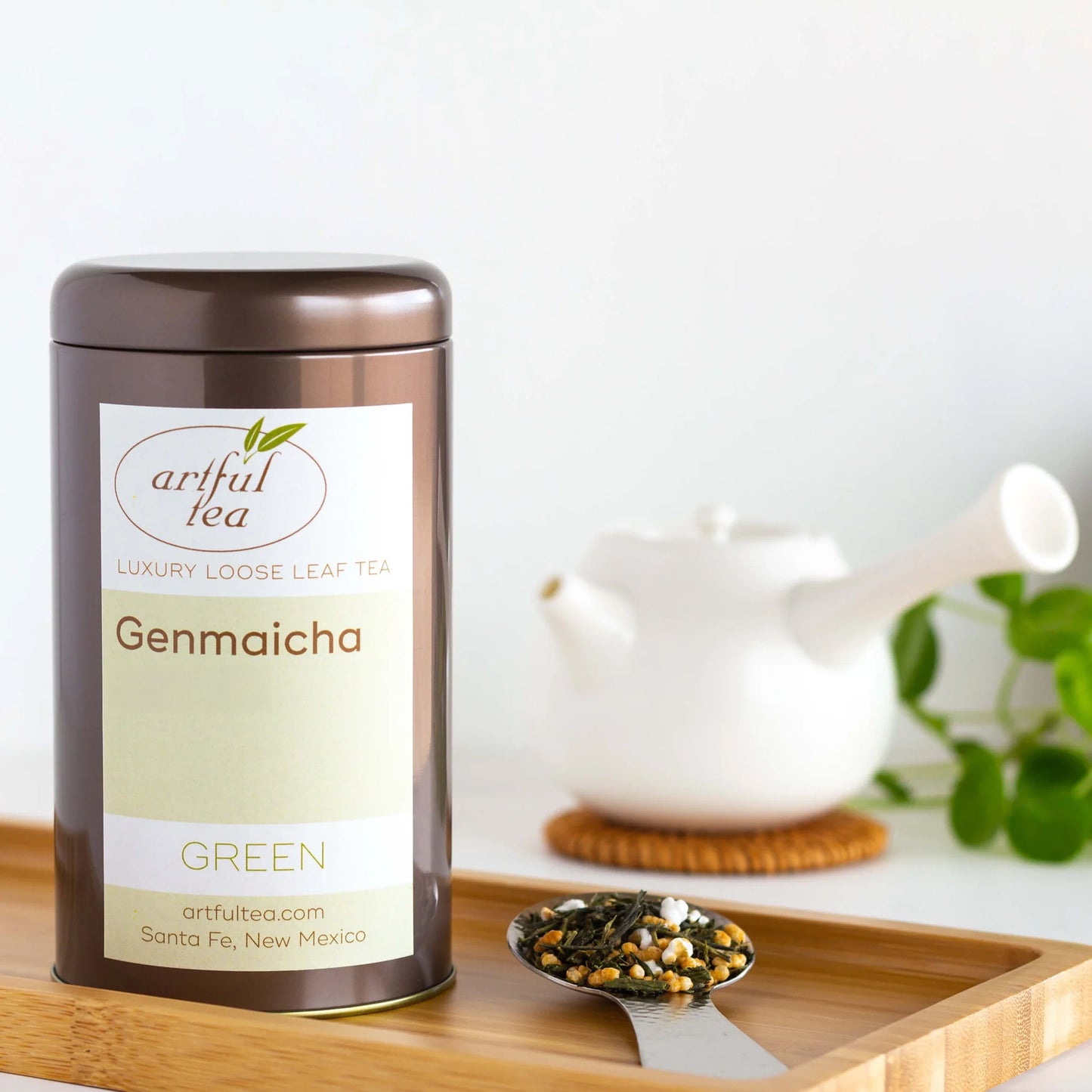Genmaicha Green Tea shown packaged in a brown tin beside a silver scoop of loose tea leaves in a rectangular, wooden tray. In the background is a white teapot on a woven coaster.