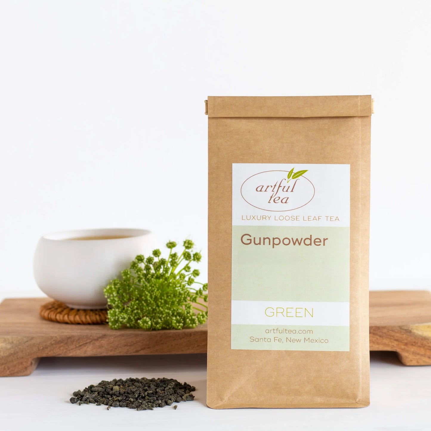 Gunpowder Green Tea shown packaged in a brown Kraft bag with loose tea leaves beside it. In the background is a white mug of brewed tea on a wooden tray.