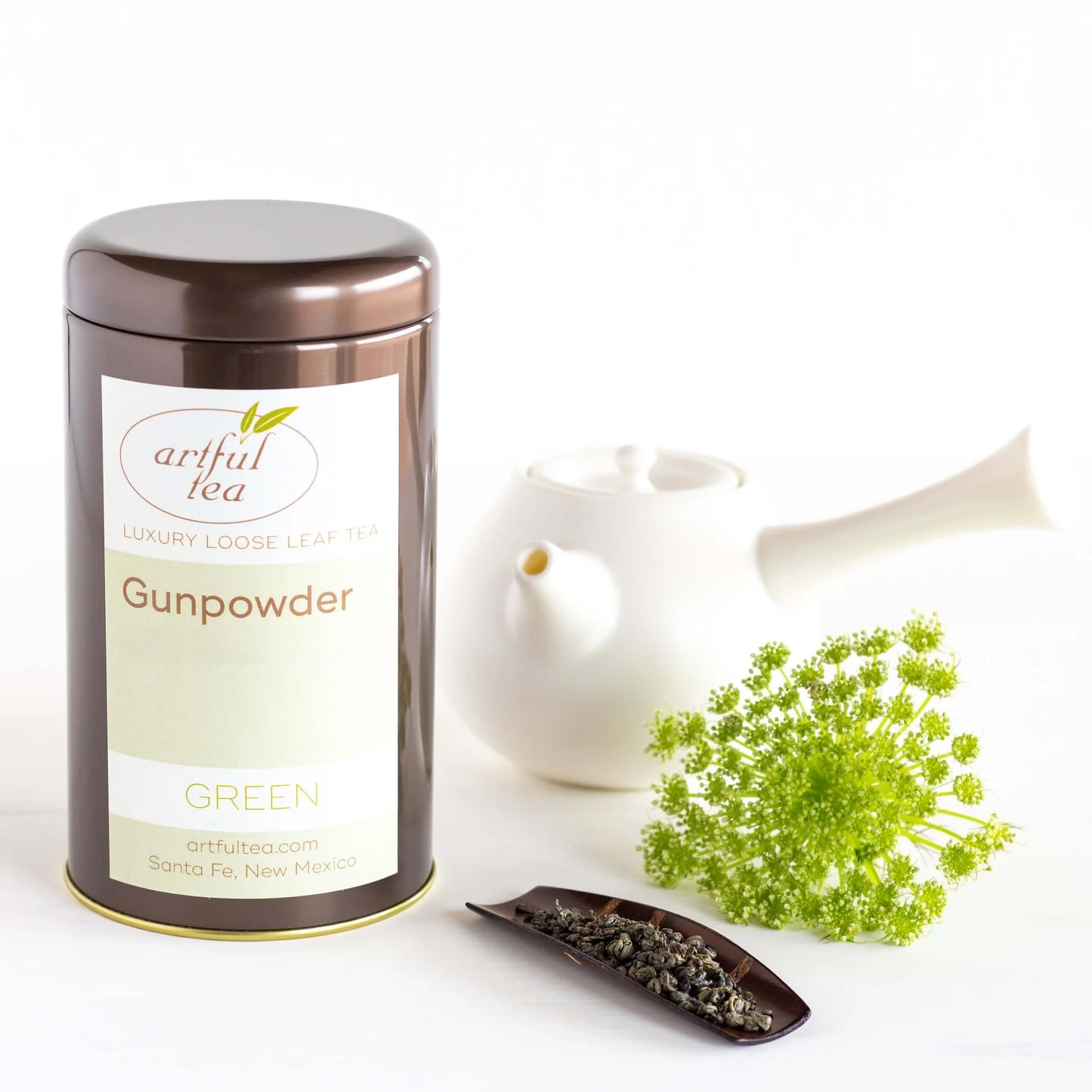 Gunpowder Green Tea shown packaged in a brown tin beside a white teapot, a plant, and brown platter full of tea leaves.