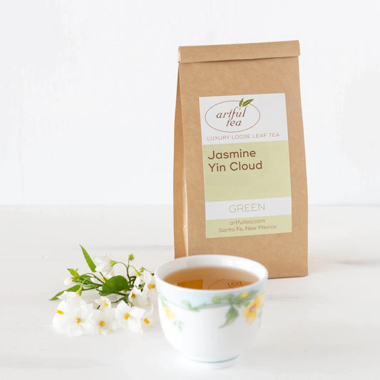 Jasmine Yin Cloud Green Tea shown packaged in a brown Kraft bag. In the foreground is a white cup with yellow flowers full of brewed tea next to a bundle of white flowers.