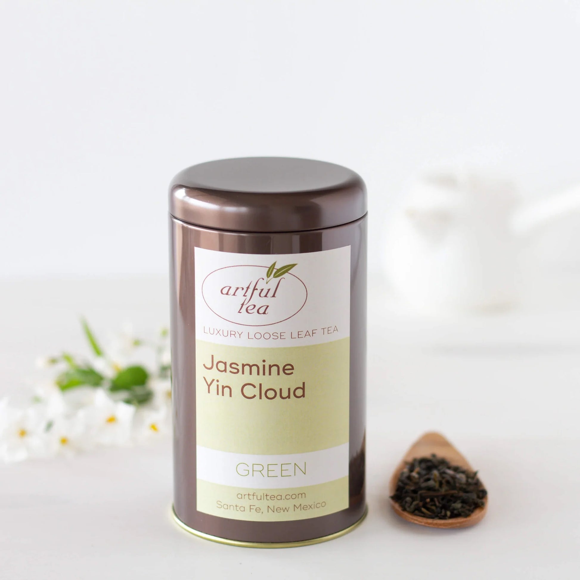 Jasmine Yin Cloud Green Tea shown packaged in a brown tin next to a small wooden scoop of loose tea leaves and a bundle of white flowers.