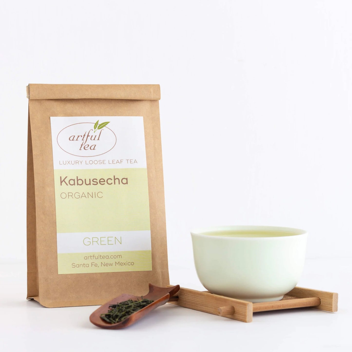Kabusecha Green Tea shown packaged in a brown Kraft bag next to a celadon teacup full of brewed tea on top of a bamboo coaster beside a wooden scoop full of tea leaves.