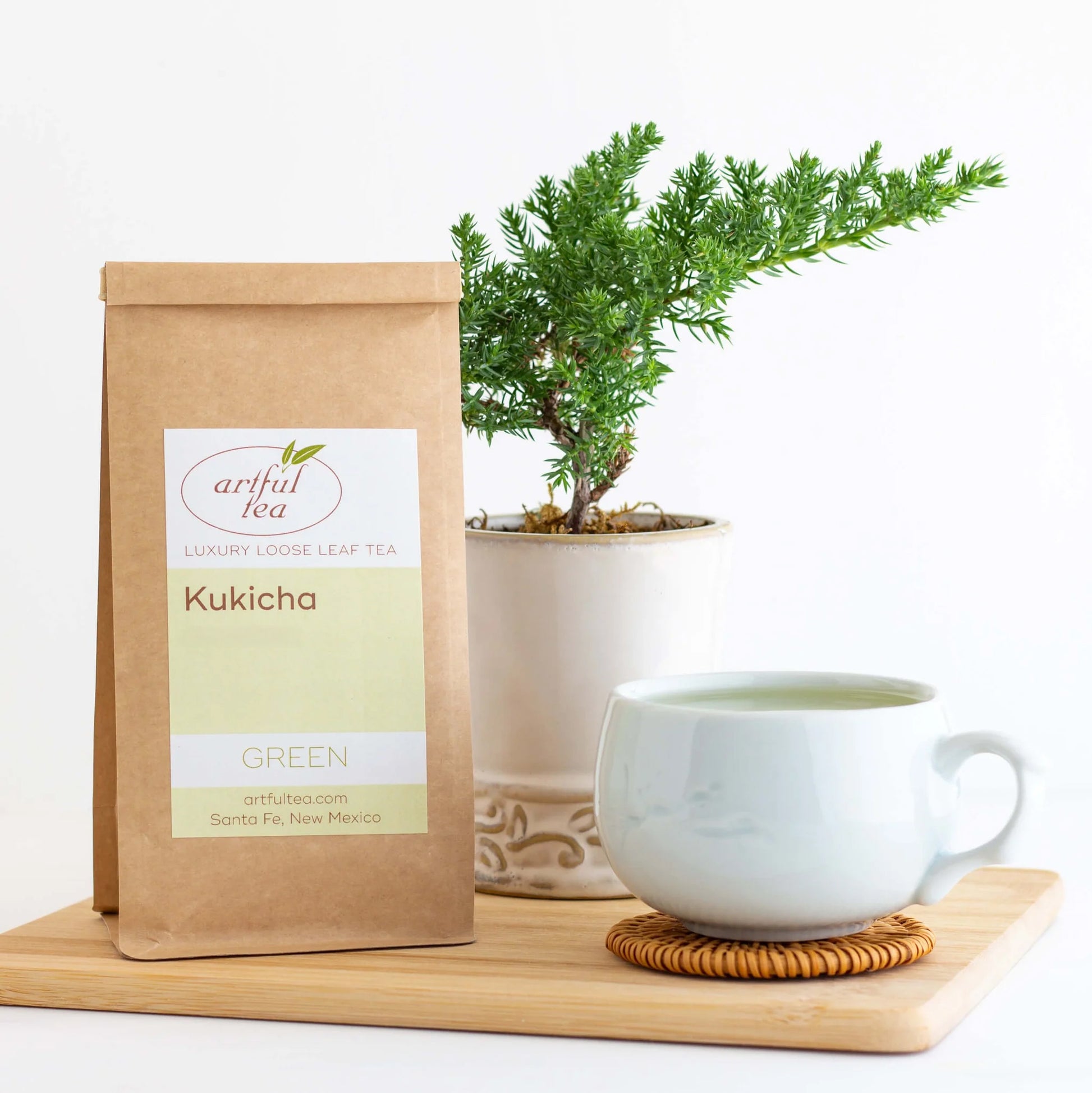 Kukicha Green Tea shown packaged in a brown Kraft bag beside a plant in a white pot and a white mug full of brewed tea on a coaster, all on top of a wooden plank.
