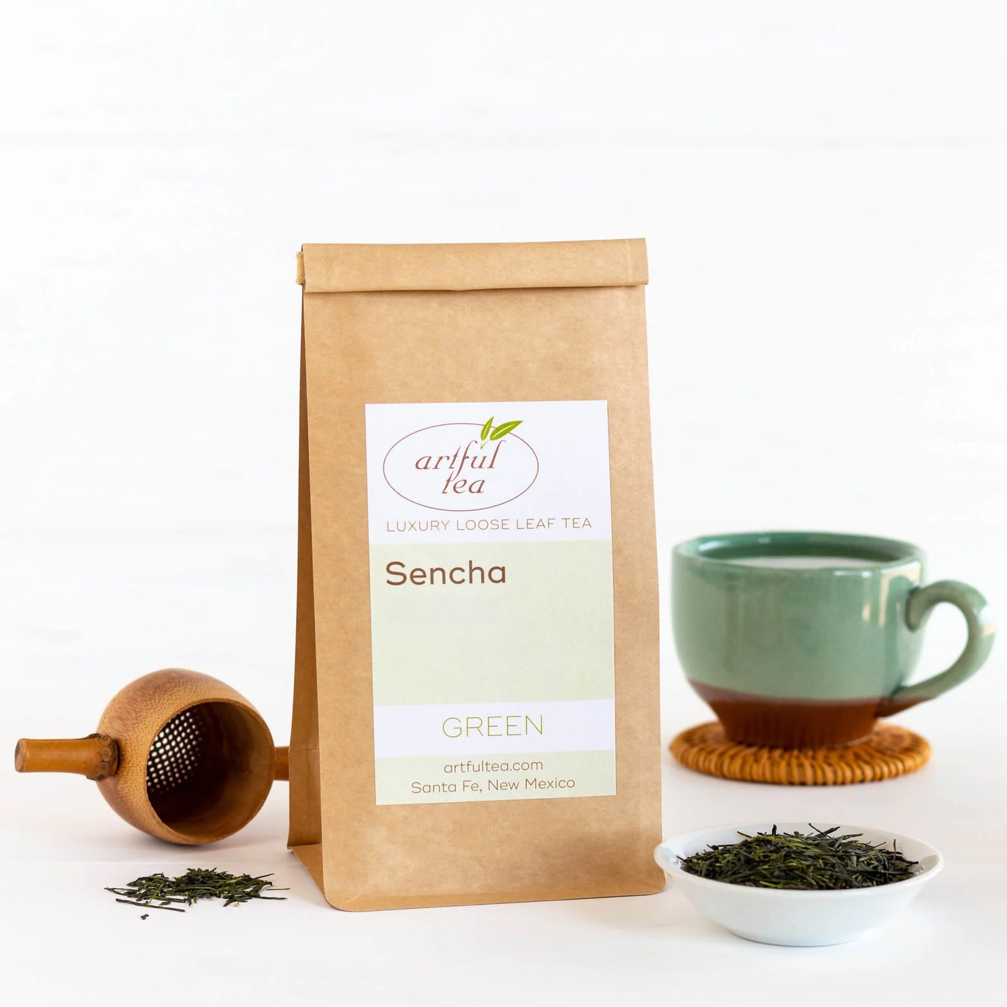 Sencha Green Tea shown packaged in a brown Kraft bag surrounded by a wooden scoop, a white bowl of loose tea leaves, and a blue and brown mug on a coaster.
