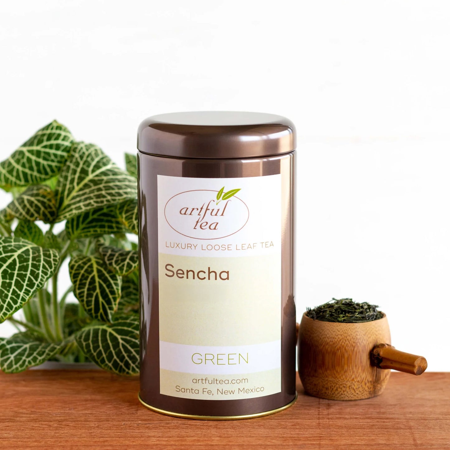 Sencha Green Tea shown packaged in a brown tin next to a wooden scoop of loose tea leaves and a plant on a wooden plank.