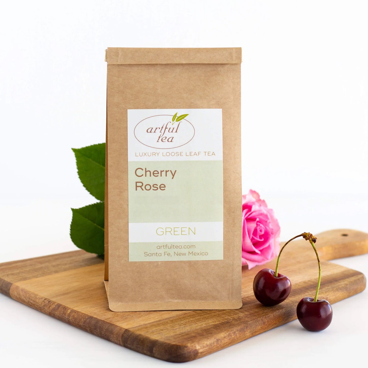 Cherry Rose Green Tea shown packaged in a brown Kraft bag on a wooden board with a pink rose and dark cherries nearby.