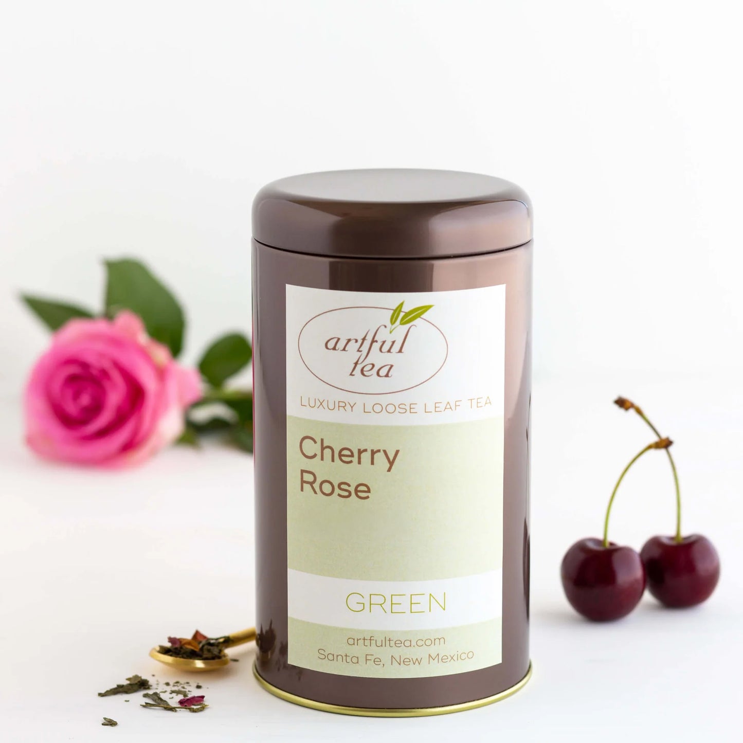 Cherry Rose Green Tea shown packaged in a brown tin with a pink rose and dark cherries nearby as well as a small golden spoon of loose tea leaves.