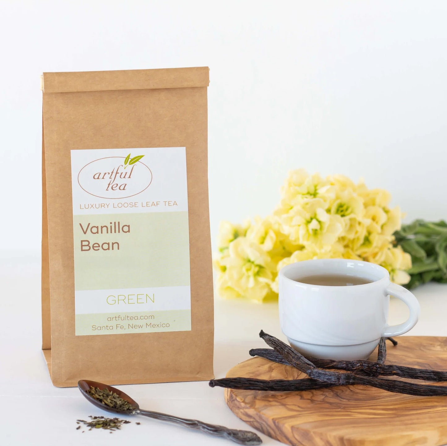 Vanilla Bean Green Tea shown packaged in a brown Kraft bag. Near it is a spoon of loose tea leaves and a white mug full of brewed tea with vanilla beans on a wooden platter. In the background is a bundle of yellow flowers.