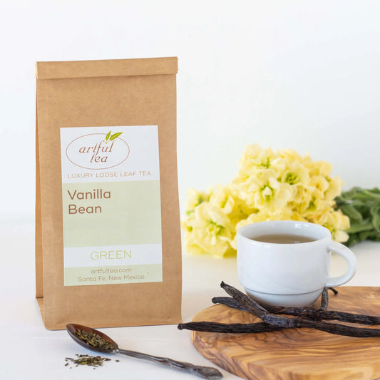 Vanilla Bean Green Tea shown packaged in a brown Kraft bag. Near it is a spoon of loose tea leaves and a white mug full of brewed tea with vanilla beans on a wooden platter. In the background is a bundle of yellow flowers.