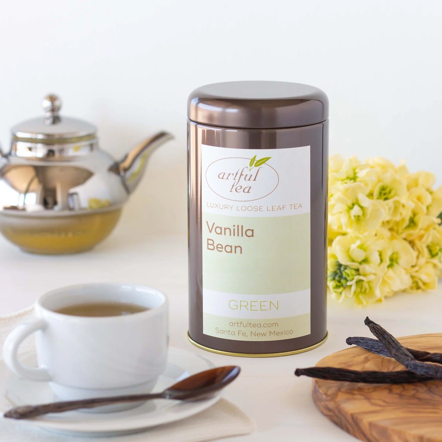 Vanilla Bean Green Tea shown packaged in a brown tin. In the foreground is a white mug full of brewed tea and a spoon on a white plate as well as vanilla beans on a wooden tray. In the background is a silver teapot and a bundle of yellow flowers.