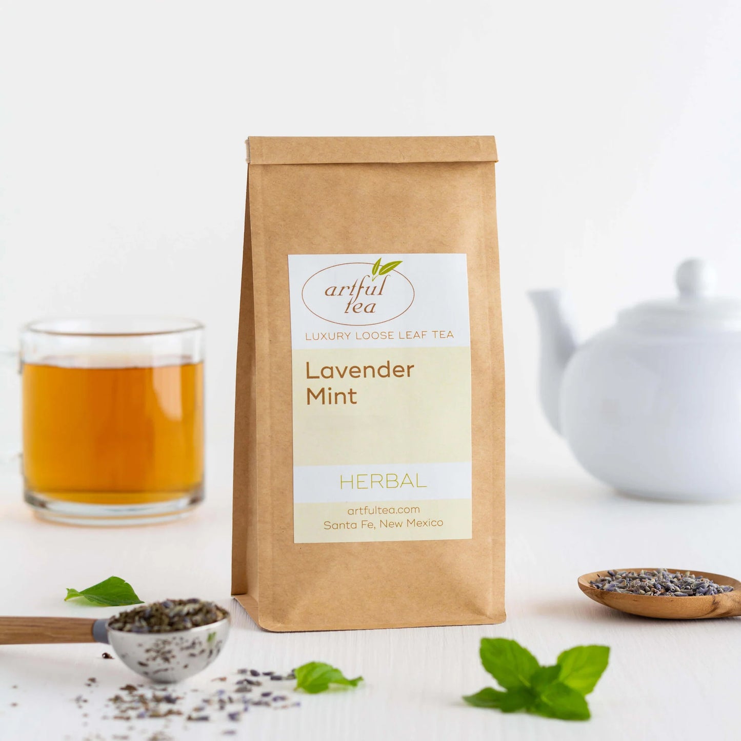 Lavender Mint Herbal Tea shown packaged in a brown Kraft bag. In the foreground is two scoops of loose tea with some mint leaves. In the background is a white teapot and a clear cup of brewed tea.
