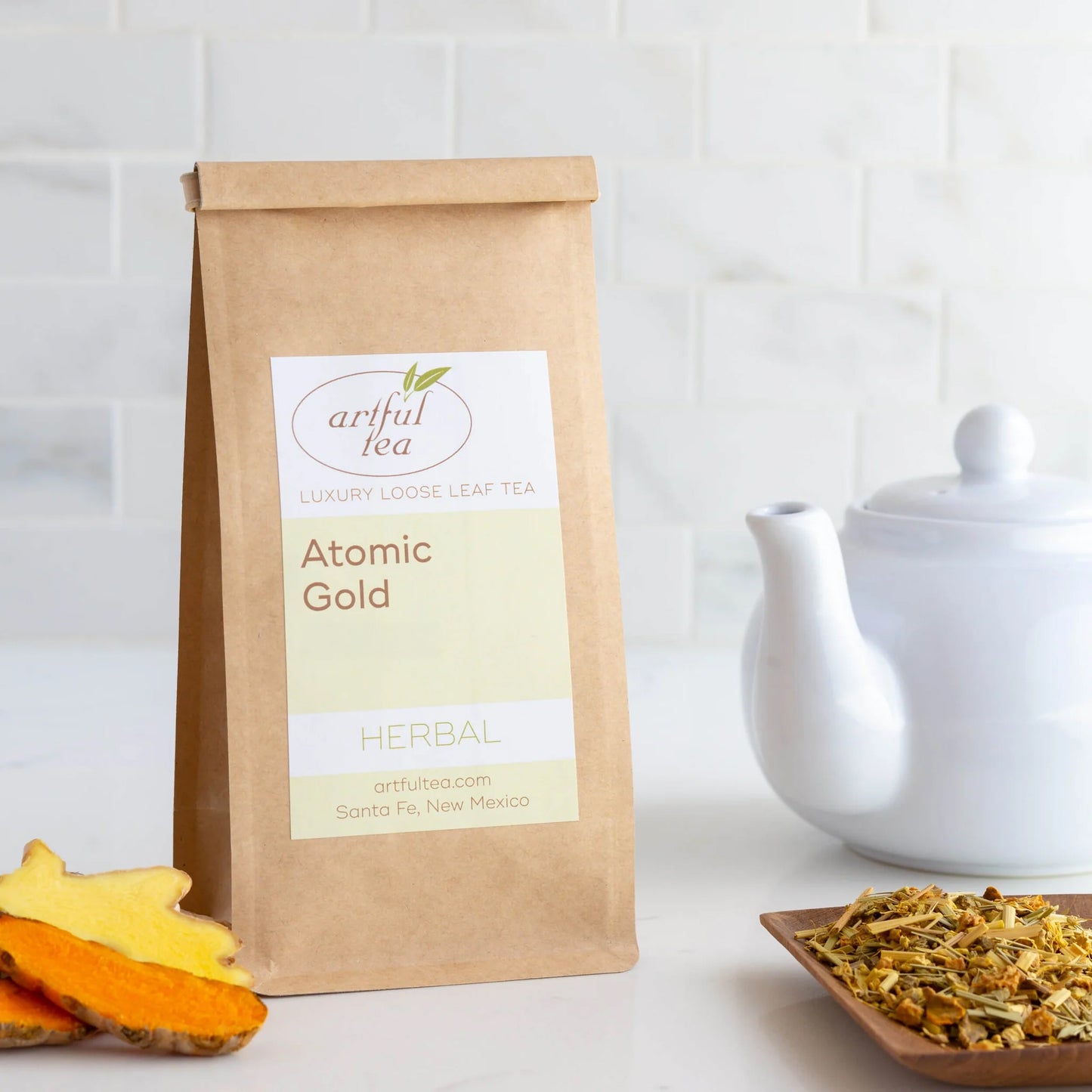 Atomic Gold Herbal Tea shown packaged in a brown Kraft bag next to a white teapot and wooden platter of loose leaf tea.