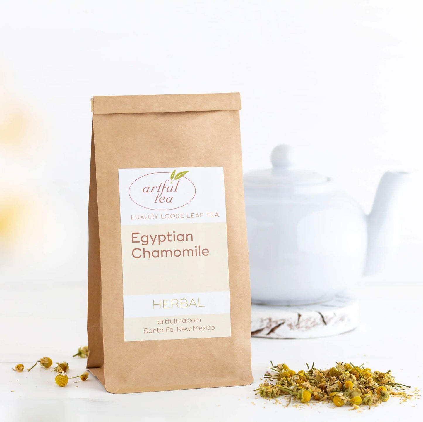 Egyptian Chamomile Herbal Tea shown packaged in a brown Kraft bag next to a pile of loose leaf tea and a white tea pot in the background.
