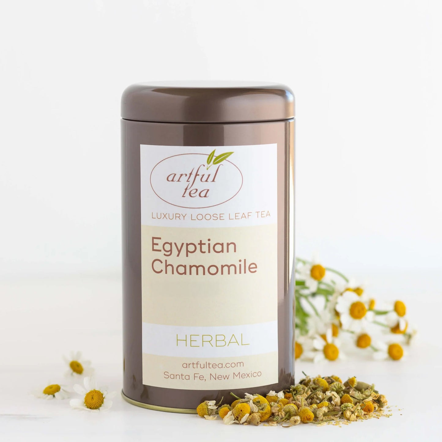 Egyptian Chamomile Herbal Tea shown packaged in a brown tin with a pile of loose leaf tea next to it and white flowers in the background.