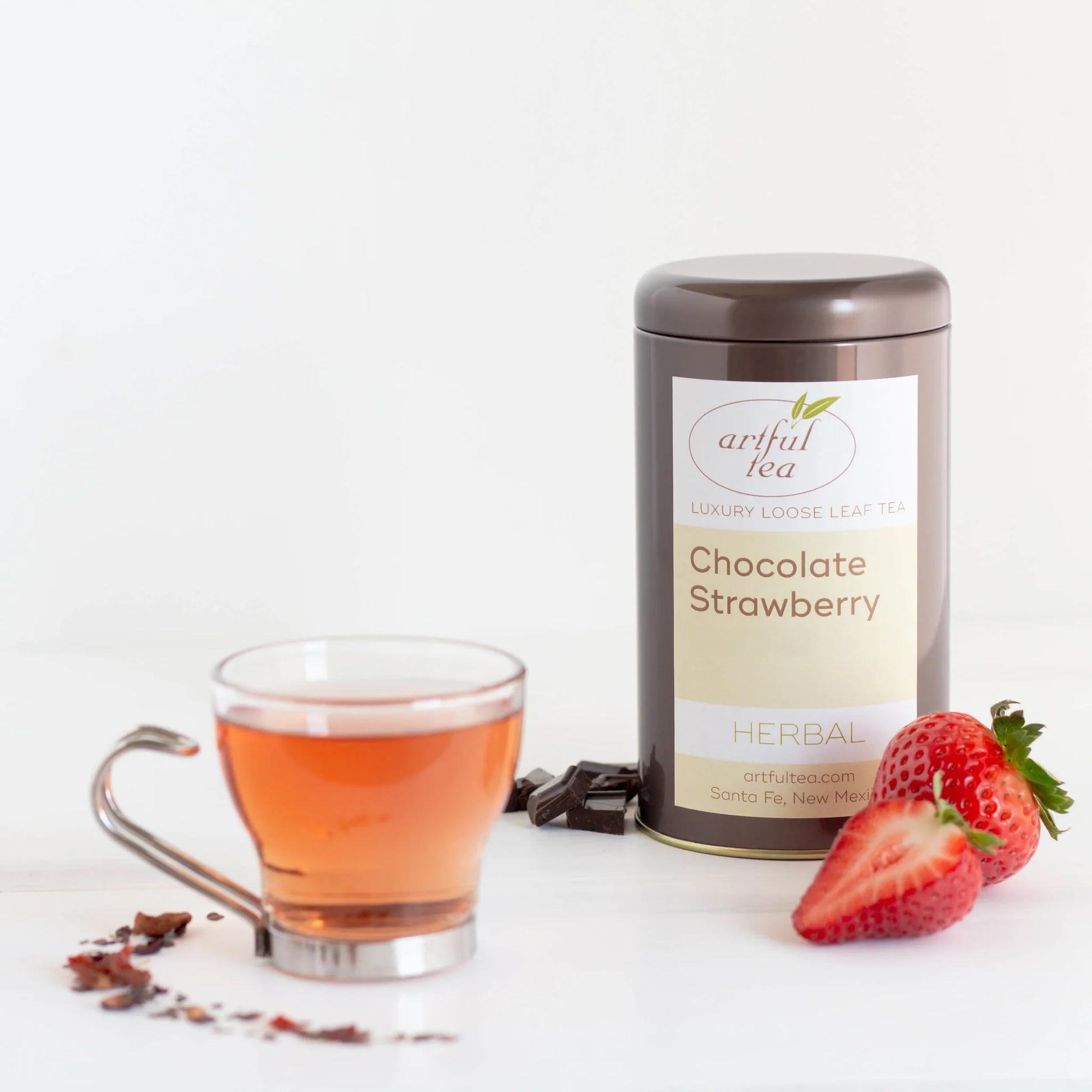 Chocolate Strawberry Herbal Tea shown packaged in a brown tin surrounded by ingredients and next to a clear cup with a metal handle full of brewed tea.