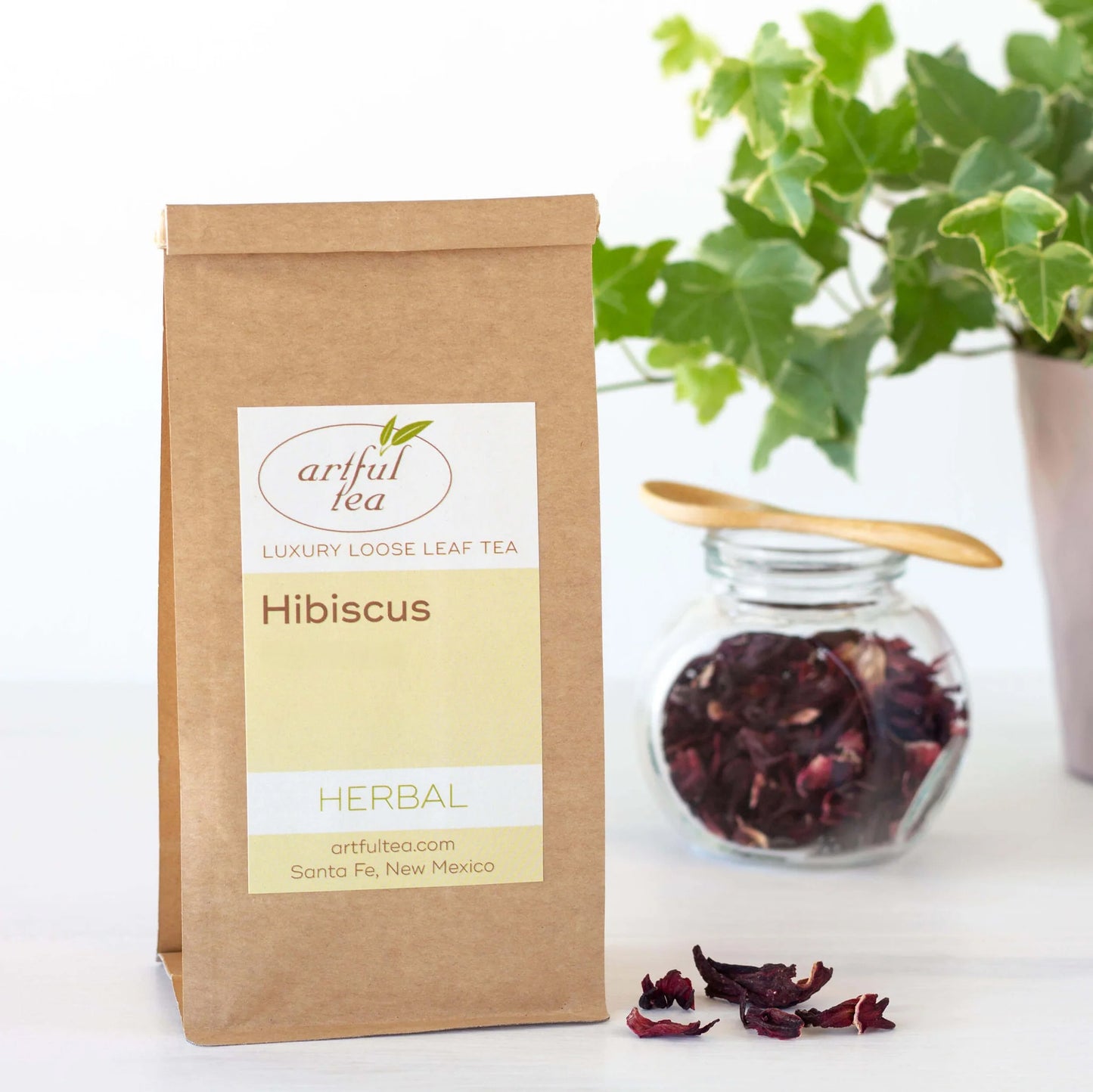 Hibiscus Herbal tea shown packaged in a brown Kraft bag next to a clear jar of loose leaf tea with a wooden spoon on top of it.