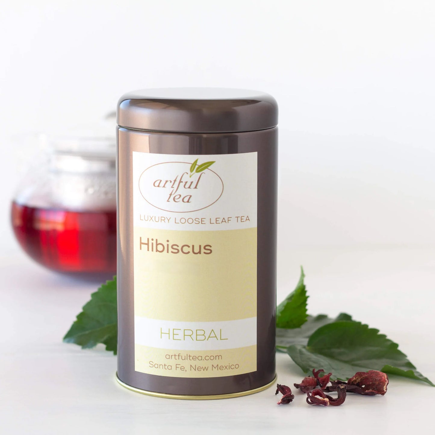 Hibiscus Herbal Tea is shown packaged in a brown tin with loose leaf tea and leaves surrounding it. In the back is a clear teapot full of brewed tea.