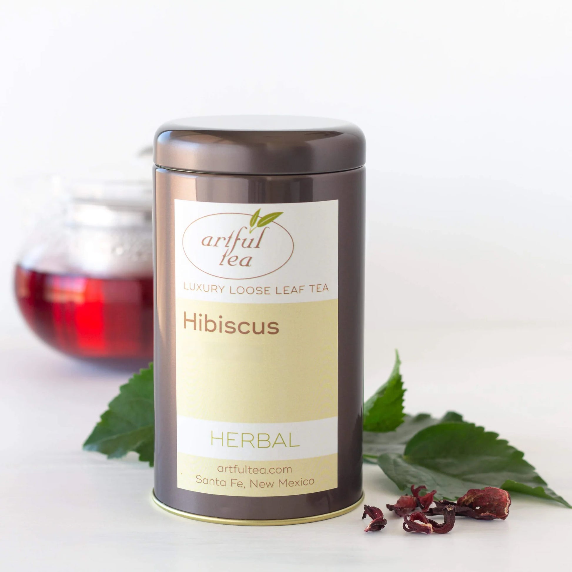 Hibiscus Herbal Tea is shown packaged in a brown tin with loose leaf tea and leaves surrounding it. In the back is a clear teapot full of brewed tea.