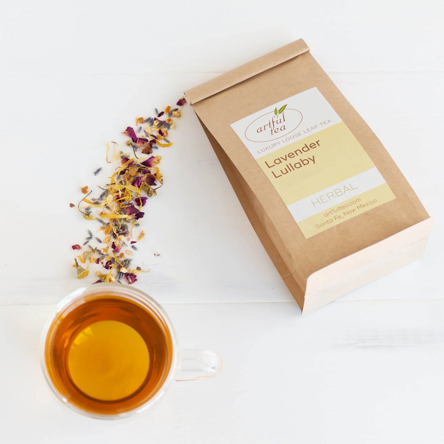 Lavender Lullaby Herbal Tea shown from above packaged in a brown Kraft bag next to a clear mug of brewed tea with loose leaf tea scattered about.