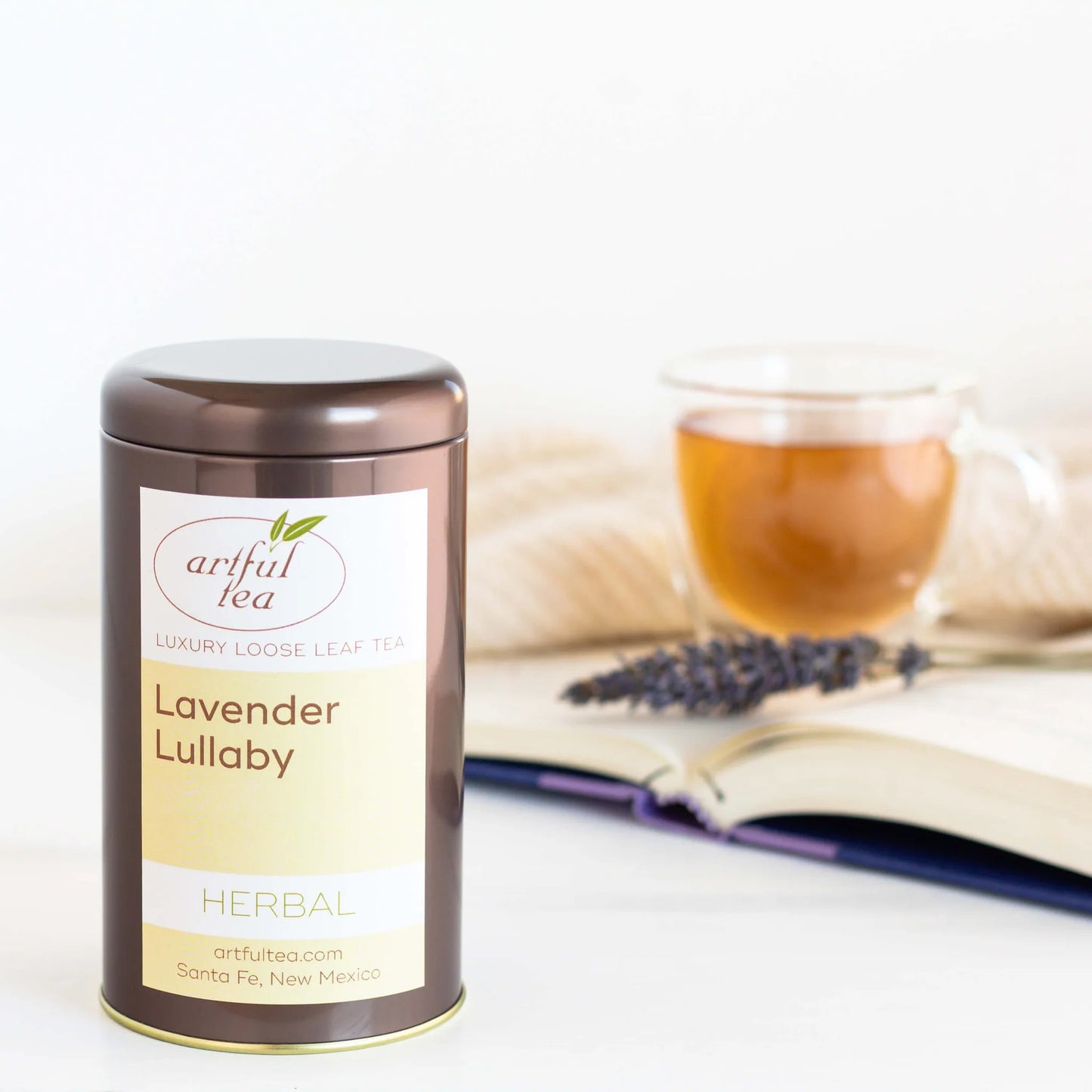 Lavender Lullaby Herbal Tea shown packaged in a brown tin. In the background is a clear mug full of brewed tea with a sprig of lavender on top of an open book.