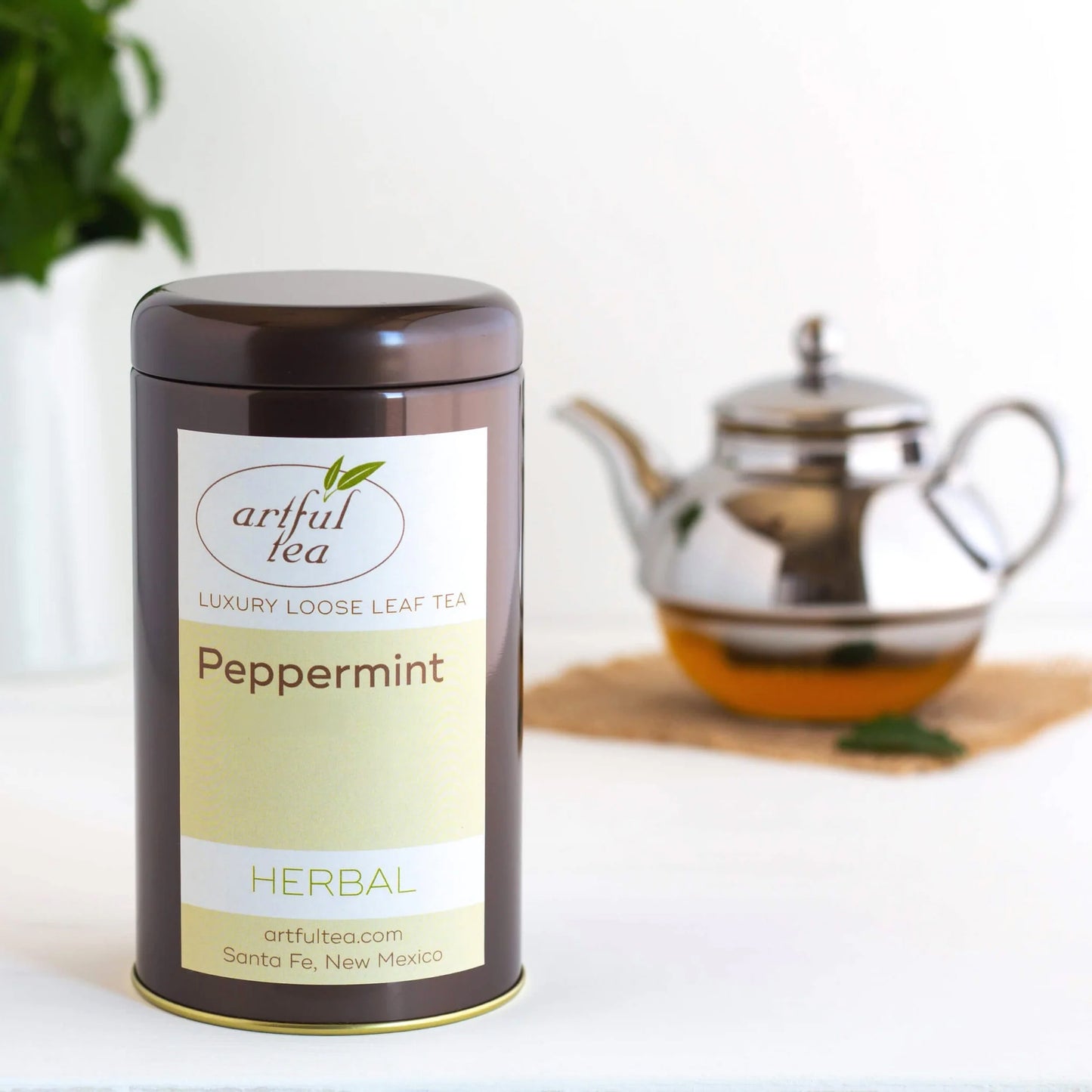 Peppermint Herbal Tea shown packaged in a brown tin. In the background is a clear teapot of brewed tea on a burlap square.