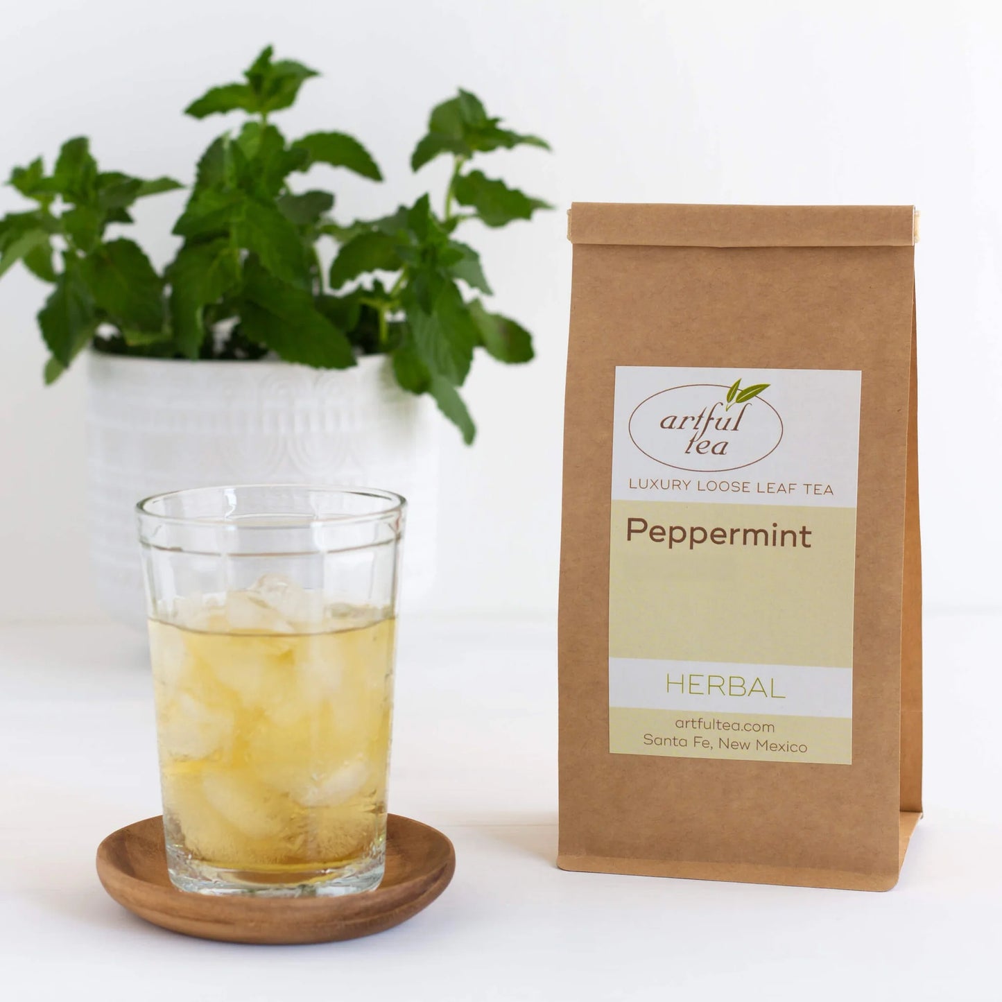 Peppermint Herbal Tea shown packaged in a brown Kraft bag next to a cup of iced tea on a wooden coaster.