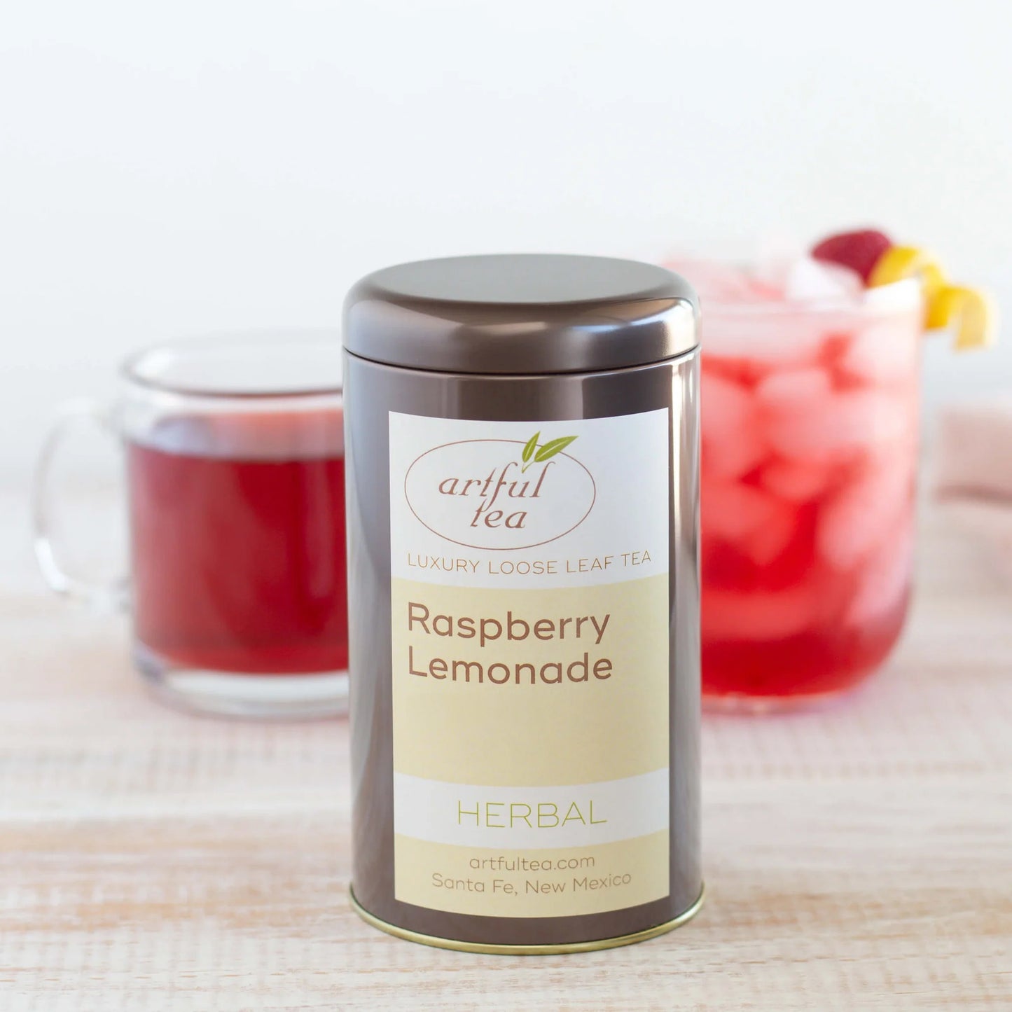 Raspberry Lemonade Herbal Tea shown packaged in a brown tin in front of a mug of hot tea and a cup of iced tea garnished with lemon and raspberries.