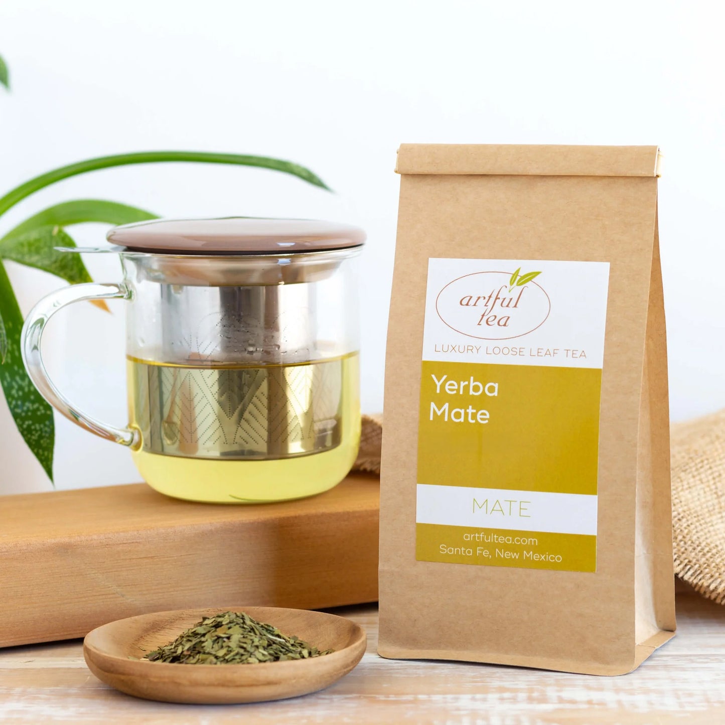 Yerba MateTea shown packaged in a brown Kraft bag next to a clear mug full of brewed tea which is on top of a wooden plank with burlap nearby. Also next to it is a small wooden bowl of loose leaf tea.