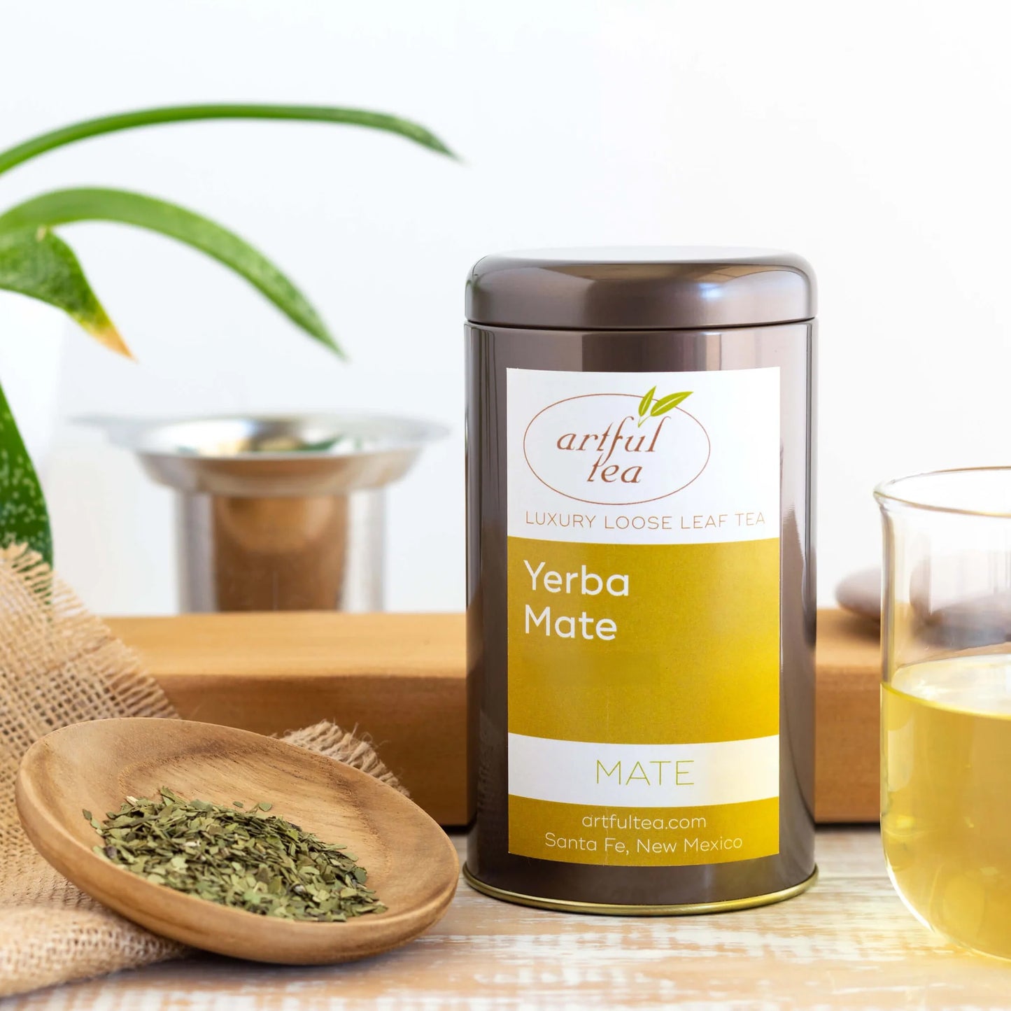Yerba Mate Tea shown packaged in a brown tin between a wooden bowl of loose leaf tea and a clear glass of brewed tea.