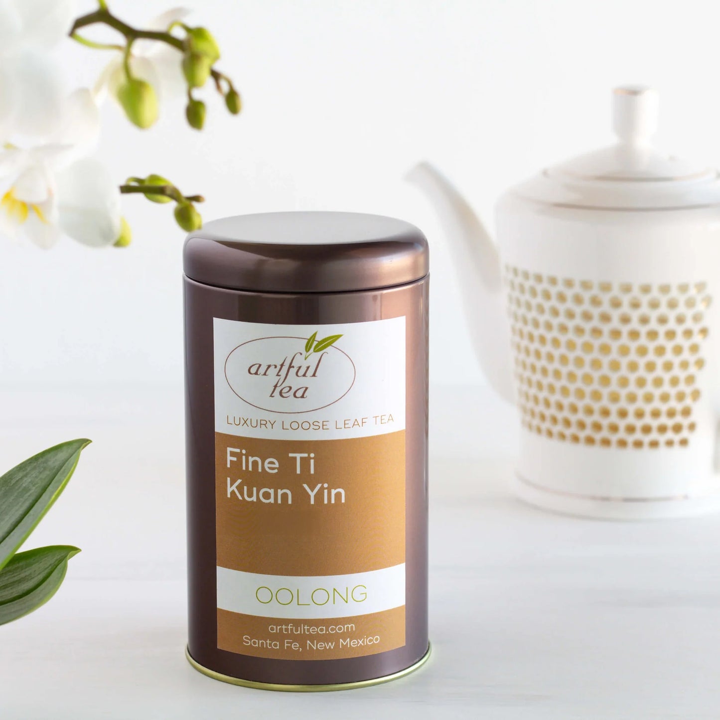Fine Ti Kuan Yin Oolong Tea shown packaged in a brown tin in between white flowers and a white teapot.