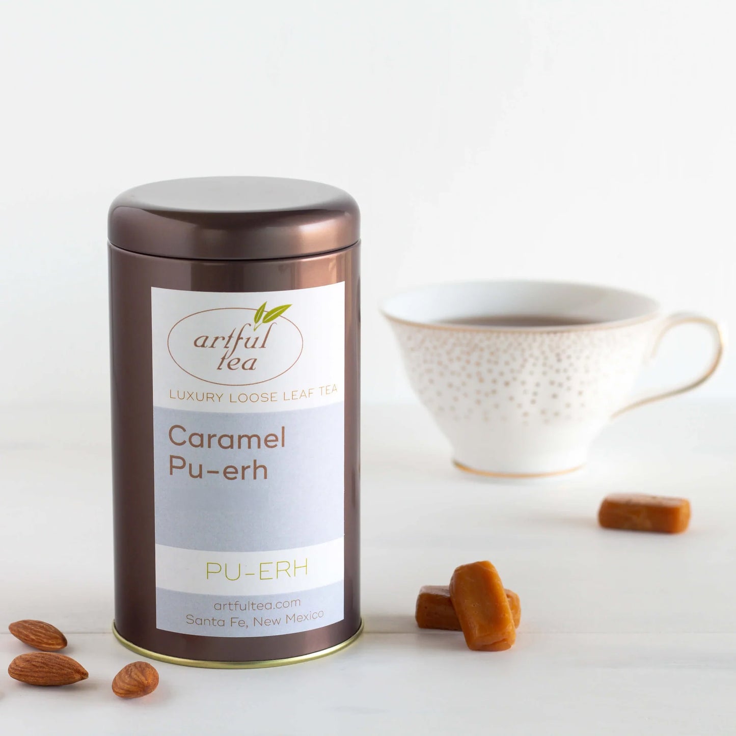 Caramel Pu-erh Tea shown packaged in a brown tin with almonds and caramel next to it. In the background is a white spotted mug full of brewed tea.