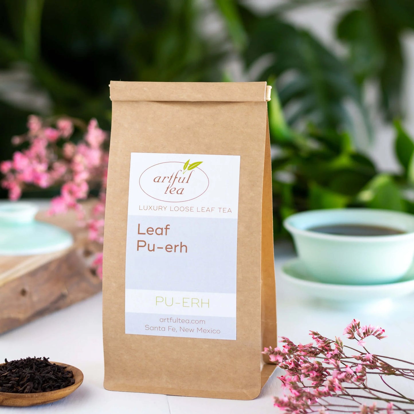 Leaf Pu-erh Tea shown packaged in a brown Kraft bag with pink flowers nearby. In the background is a blue celadon gaiwan cup and in the foreground is a wooden spoon of loose leaf tea.