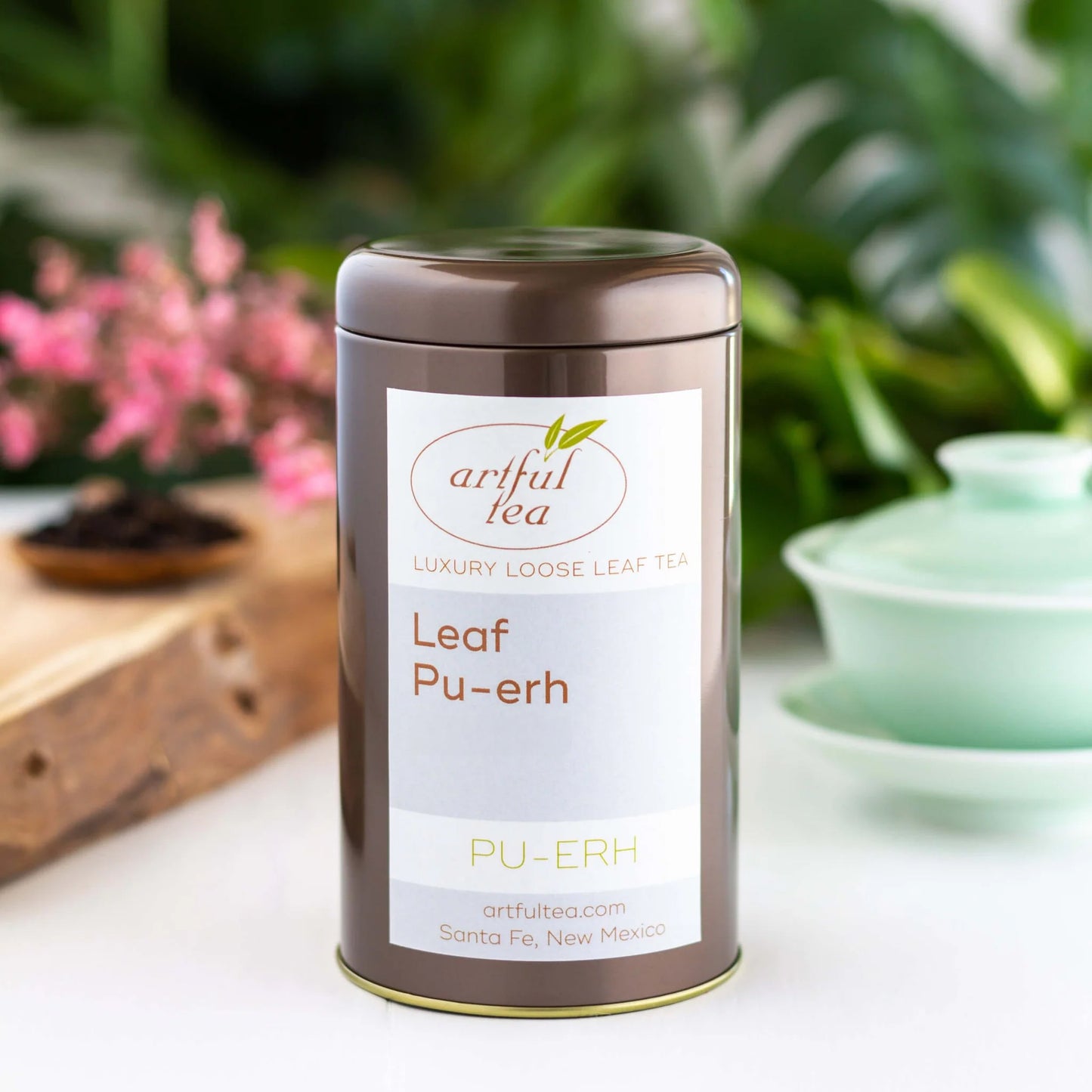 Leaf Pu-erh Tea shown packaged in a brown tin. In the background are pink flowers on a wooden plank and a blue gaiwan cup.