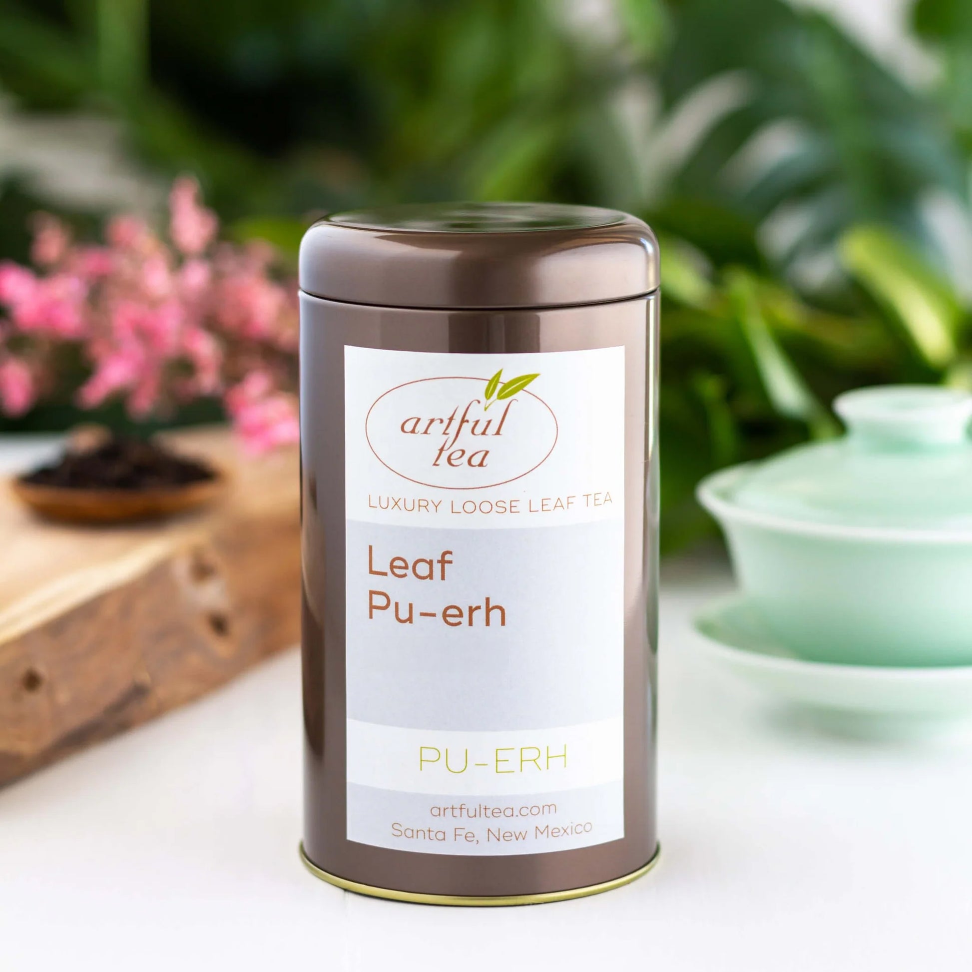 Leaf Pu-erh Tea shown packaged in a brown tin. In the background are pink flowers on a wooden plank and a blue gaiwan cup.