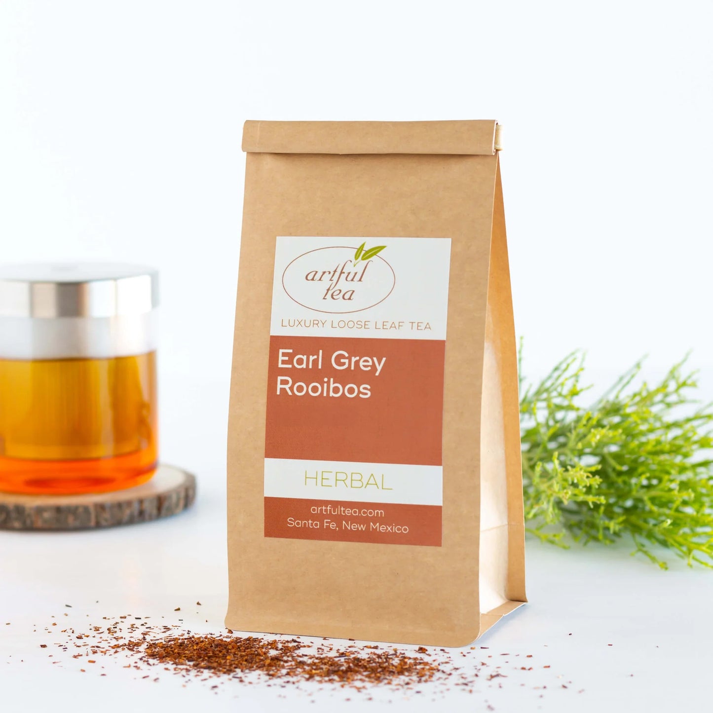 Earl Grey Rooibos Herbal Tea shown packaged in a brown Kraft bag next to a clear cup of brewed tea and tree sprigs.