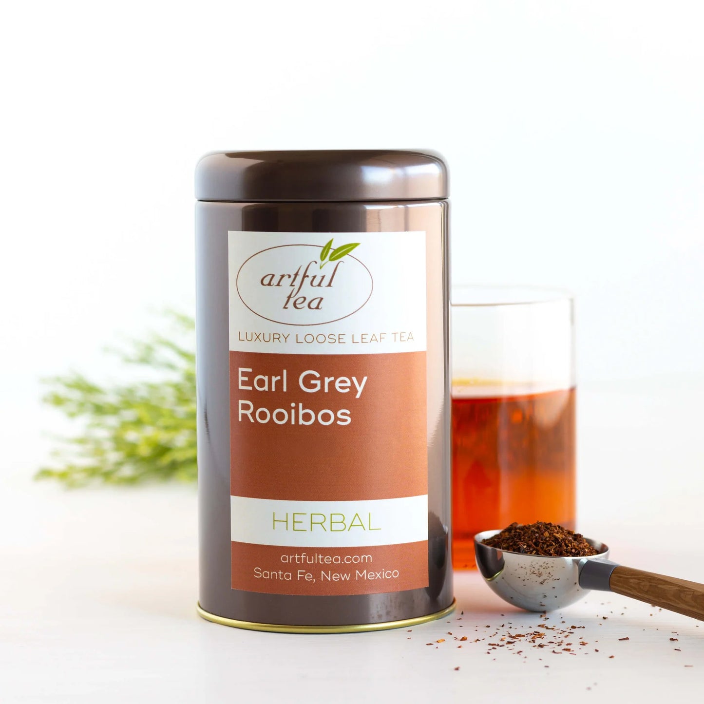 Earl Grey Rooibos Herbal Tea shown packaged in a brown tin next to a clear cup of brewed tea and a scoop of loose leaf tea.