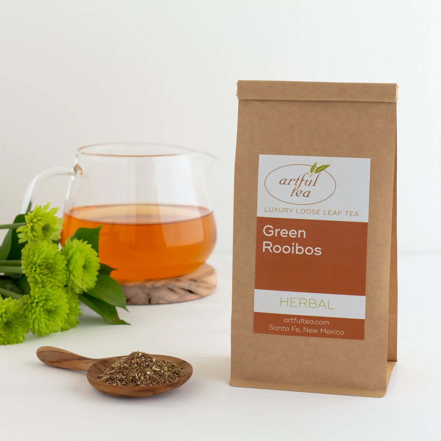Green Rooibos Herbal Tea shown packaged in a brown Kraft bag next to a wooden scoop of loose leaf tea. In the background is a clear mug full of brewed tea next to green flowers.