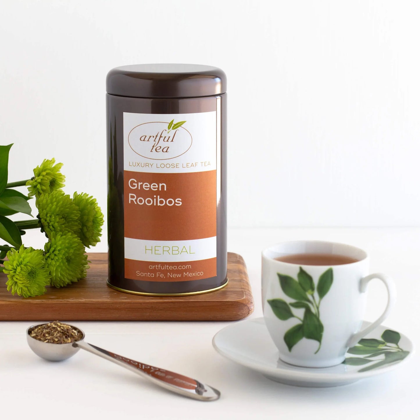 Green Rooibos Herbal Tea shown packaged in a brown tin next to green flowers on top of a wooden tray. In the foreground is a teaspoon of loose leaf tea and a white mug full of brewed tea.