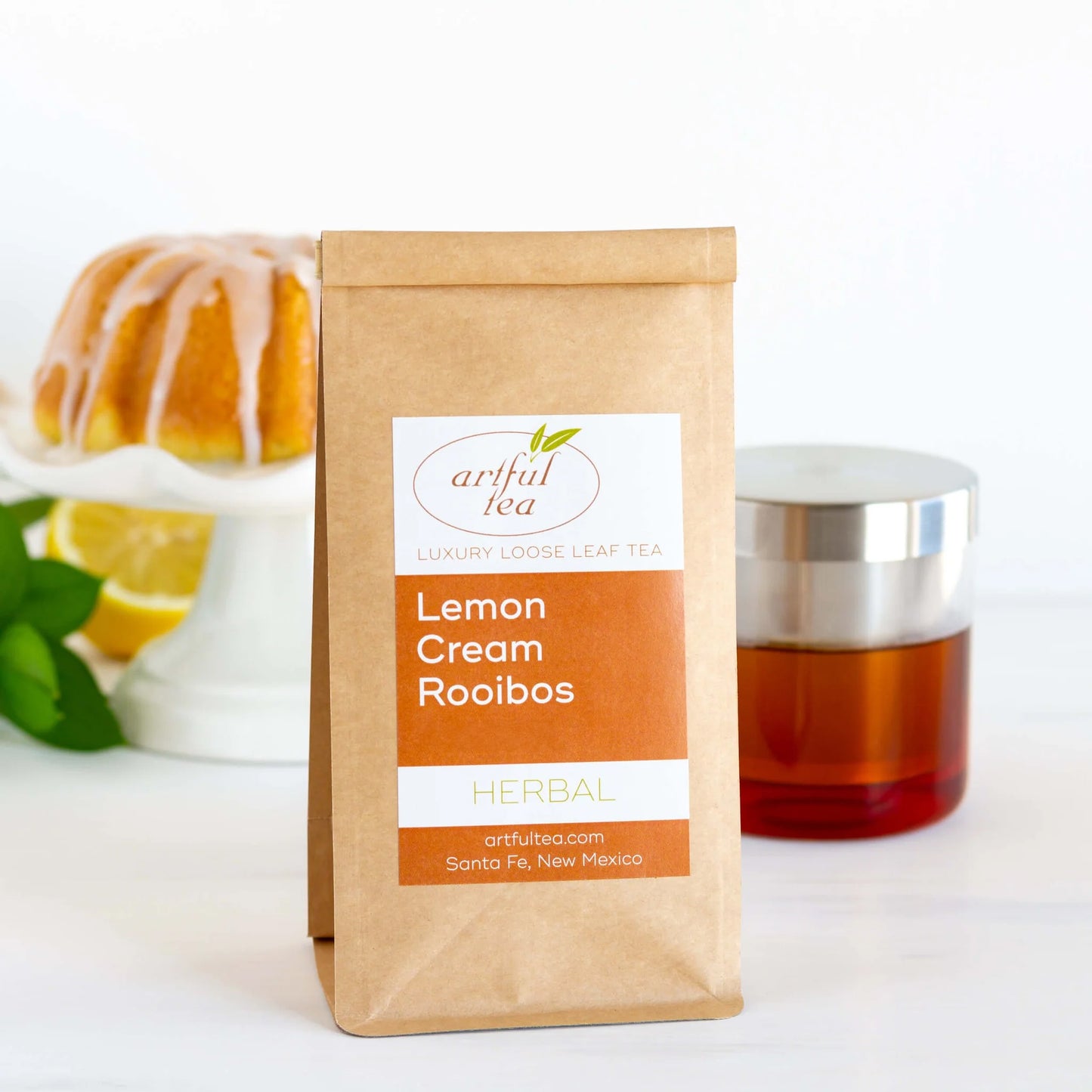 Lemon Cream Rooibos Herbal Tea shown packaged in a brown Kraft bag in front of a clear cup of brewed tea and a cake on a white platter.