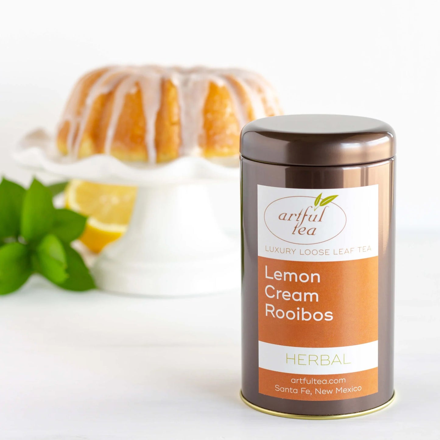 Lemon Cream Rooibos Herbal Tea shown packaged in a brown tin. In the background is a white platter with a cake on it.